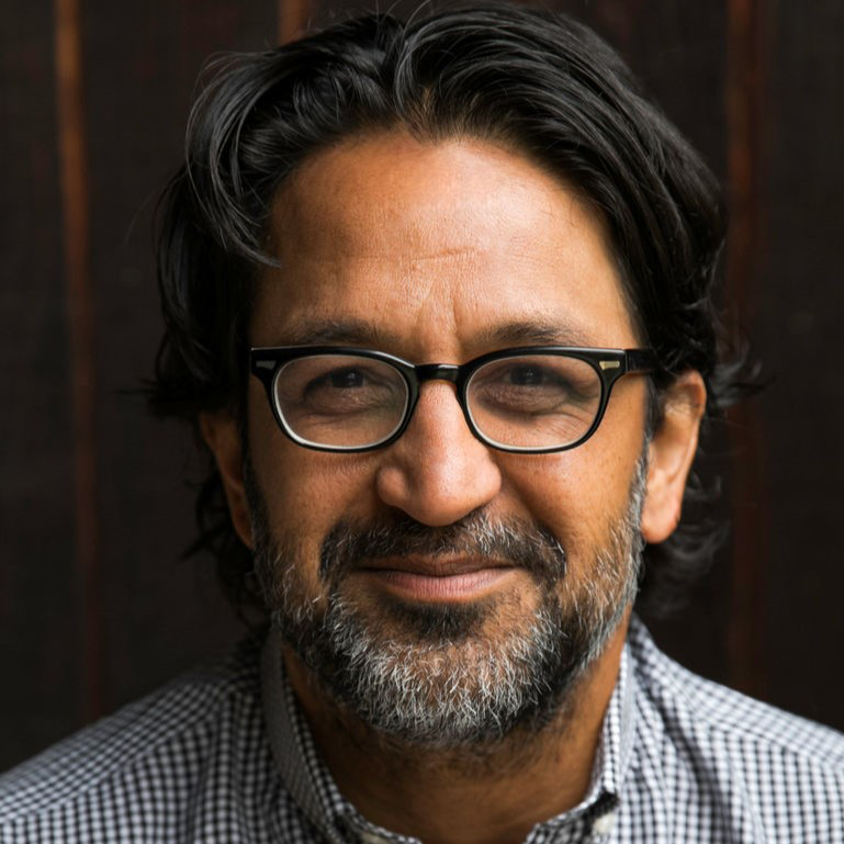 Headshot of Lauren Francis-Sharma, a smiling Black woman wearing glasses, a patterned scarf, and a white top